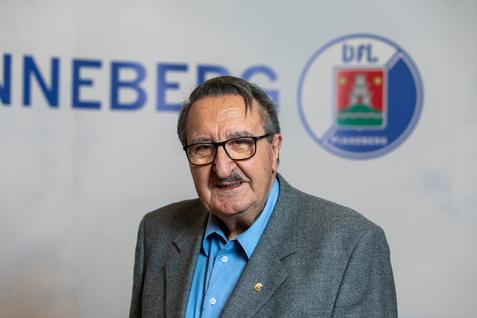 Porträt eines älteren Mannes mit Brille, grauem Blazer und blauer Hemd vor einem unscharfen Hintergrund mit Schriftzug.