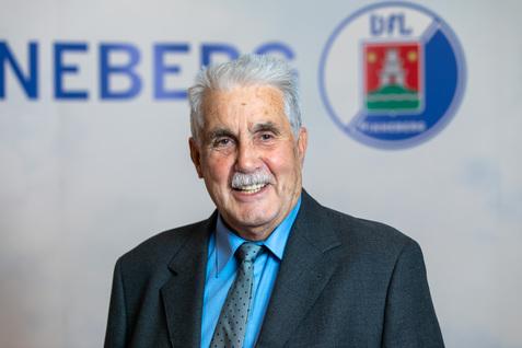 Lächelnder älterer Mann in Anzug und Krawatte vor einem bunten Hintergrund mit Logos und Schriftzügen.
