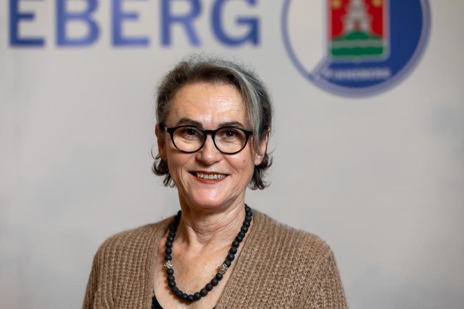 Porträt einer Frau mit Brille und grauen Strähnen, die in einem gemütlichen Pullover und einer Halskette lächelt.