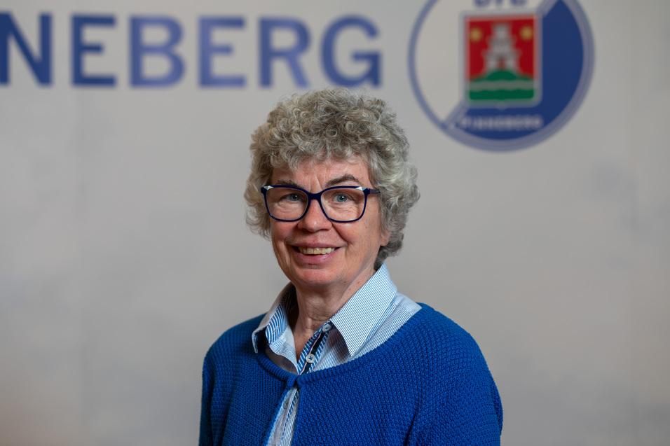 Porträt einer lächelnden Frau mit lockigem, grauem Hair und blauem Pullover, vor einem neutralen Hintergrund mit Schriftzug.