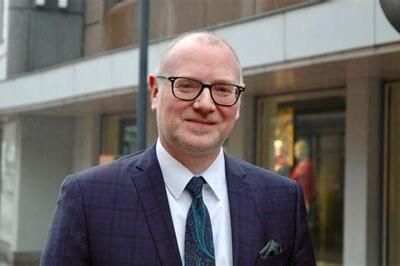 Mann in Anzug mit Brille steht auf einer Stra&szlig;e vor Schaufenstern, l&auml;chelt und tr&auml;gt eine bunte Krawatte.