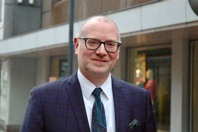 Mann in Anzug mit Brille steht auf einer Stra&szlig;e vor Schaufenstern, l&auml;chelt und tr&auml;gt eine bunte Krawatte.