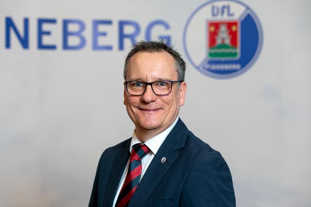 Porträt eines Mannes mit Brille und Anzug, lächelnd vor einem Hintergrund mit dem Schriftzug "NEBERG".