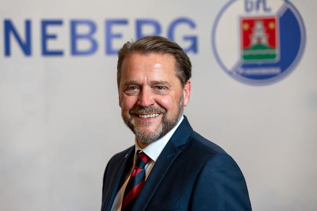 Porträt eines lächelnden Mannes in einem blauen Anzug mit gestreifter Krawatte vor einem unscharfen Hintergrund.