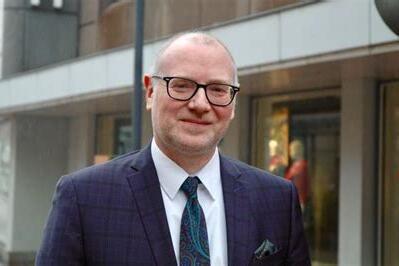 Mann in Anzug mit Brille steht auf einer Straße vor Schaufenstern, lächelt und trägt eine bunte Krawatte.