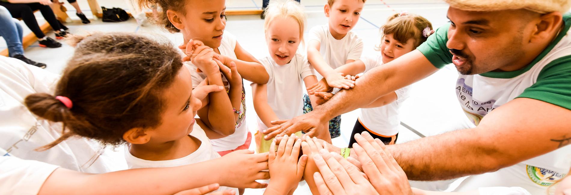 Gruppenspiel mit Kindern, die ihre Hände zum gemeinsamen Aufeinandertreffen heben, unter Anleitung eines Erwachsenen.