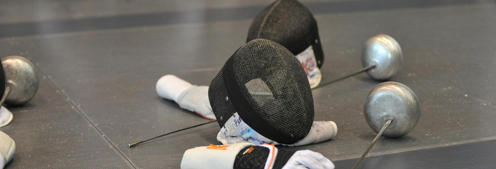 Zwei Fechthandschuhe und Masken liegen auf einer Matte, daneben stehen metallische Fechtwaffen.