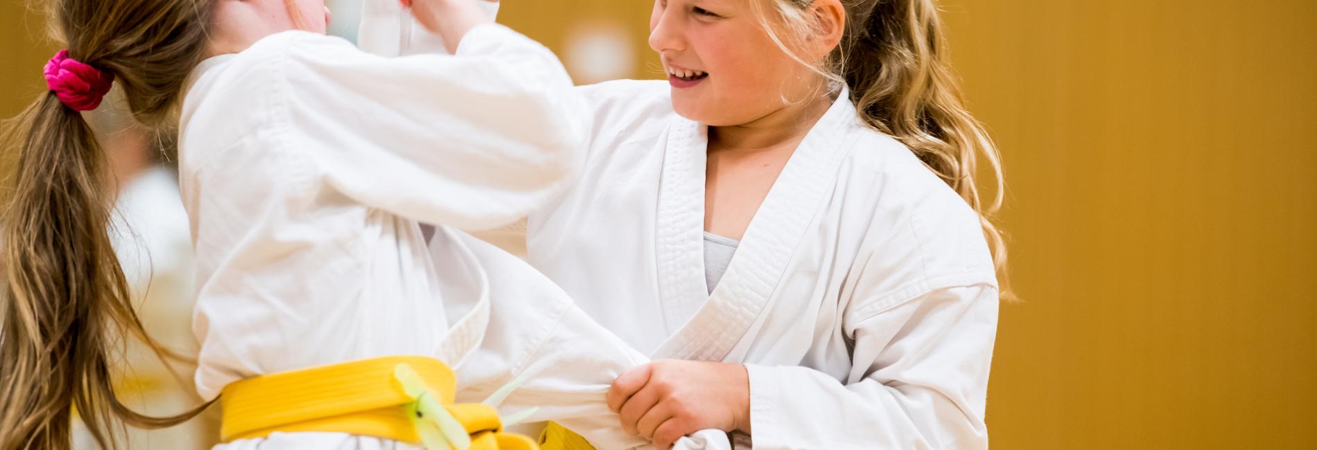 Zwei Mädchen in weißen Karateanzügen mit gelben Gurten üben freundschaftlich im Trainingsraum miteinander.