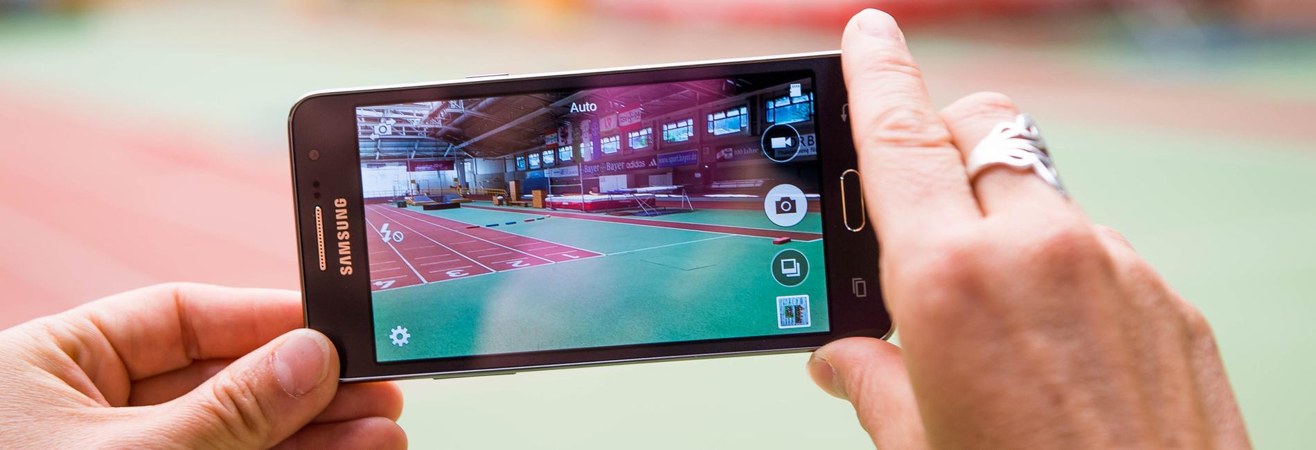 Hand hält ein Smartphone, das eine Sporthalle mit einer Laufbahn im Hintergrund zeigt.